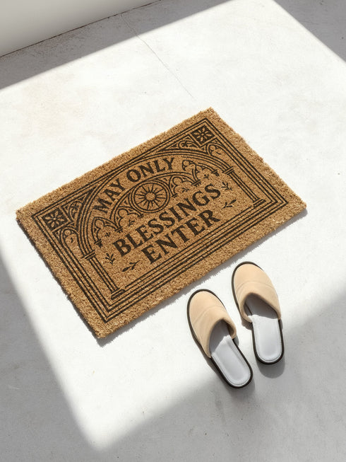 A rectangular natural brown coir doormat with a rugged texture and a durable black vinyl backing. The doormat features an intricate black design framed by a gothic arch with stylized quatrefoil details in the corners. Inside the arch, the words "MAY ONLY BLESSINGS ENTER" are printed in an elegant, bold serif font. A central sunburst medallion with radiating lines sits above the text, reinforcing the spiritual and protective theme of this gothic entryway welcome mat.