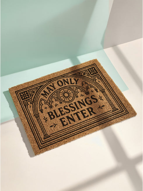 A rectangular natural brown coir doormat with a rugged texture and a durable black vinyl backing. The doormat features an intricate black design framed by a gothic arch with stylized quatrefoil details in the corners. Inside the arch, the words "MAY ONLY BLESSINGS ENTER" are printed in an elegant, bold serif font. A central sunburst medallion with radiating lines sits above the text, reinforcing the spiritual and protective theme of this gothic entryway welcome mat.