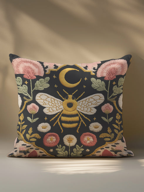 A square black pillow cover made from a pre-shrunk polyester fabric with a soft, linen-like texture. The double-sided, high-fidelity print features a large, central bee with intricate gold and white faux-embroidery on its wings and striped body. Directly above the bee is a golden crescent moon with small stars. The central design is framed by a symmetrical border of botanical elements, including pink thistle-like flowers, white and red daisies, and green leaves.