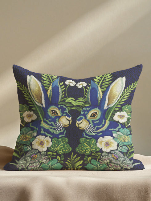 A Grimoire and Goblet decorative throw pillow cover featuring a high-fidelity print of two symmetrical hares facing each other. The design mimics the texture of raised embroidery with faux-stitches in antique gold, ochre, and royal blue threads against a deep midnight indigo background. The rabbits are surrounded by a lush wreath of green ferns and cream primrose flowers in a vintage medallion style. The fabric has a smooth, linen-like texture, visible as a soft matte finish