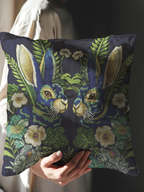 A Grimoire and Goblet decorative throw pillow cover featuring a high-fidelity print of two symmetrical hares facing each other. The design mimics the texture of raised embroidery with faux-stitches in antique gold, ochre, and royal blue threads against a deep midnight indigo background. The rabbits are surrounded by a lush wreath of green ferns and cream primrose flowers in a vintage medallion style. The fabric has a smooth, linen-like texture, visible as a soft matte finish