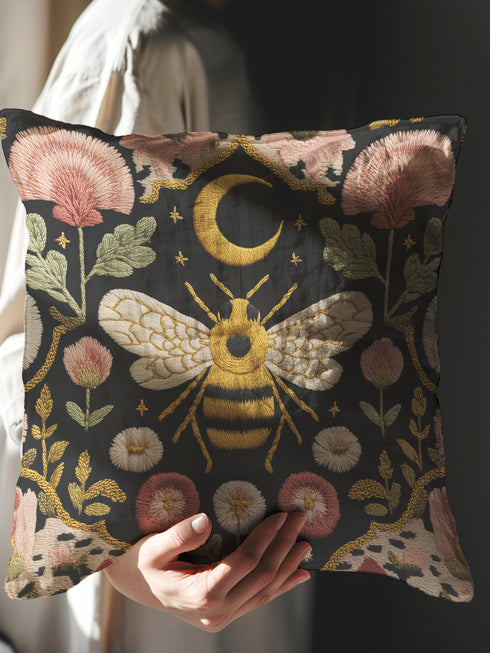 A square black pillow cover made from a pre-shrunk polyester fabric with a soft, linen-like texture. The double-sided, high-fidelity print features a large, central bee with intricate gold and white faux-embroidery on its wings and striped body. Directly above the bee is a golden crescent moon with small stars. The central design is framed by a symmetrical border of botanical elements, including pink thistle-like flowers, white and red daisies, and green leaves.
