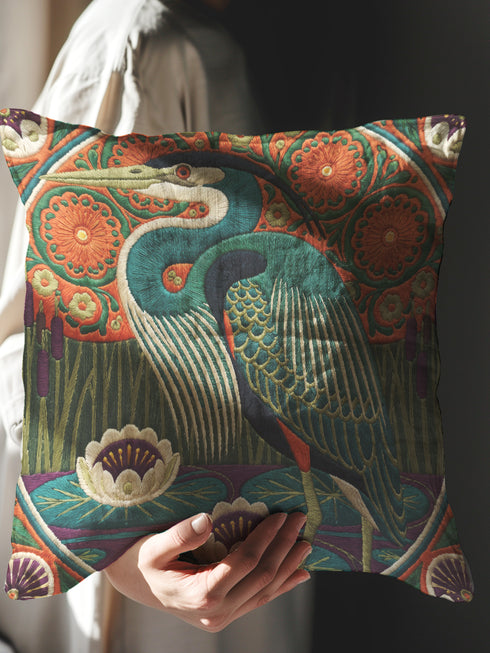A square decorative pillowcase printed with a smooth, tapestry-like design. The central image is a large Great Blue Heron in profile, rendered in shades of teal, navy, and cream. The bird stands among stylized water lilies with cream petals and mustard centers, resting on deep plum and green waters. The background features tall green grass, purple cattails, and large, sun-like circular orange floral medallions. The detailed, embroidered-style pattern uses rich, earthy, maximalist colors.