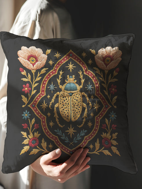 A square, dark charcoal pillow cover with a linen-feel texture, featuring an intricate, embroidered-style design. At the center, a large Egyptian scarab beetle in gold and turquoise is framed by a red and gold quatrefoil border. Symmetrical blush-pink poppies and smaller blue and red flowers with golden foliage flank the central motif, creating a lush botanical scene. Small celestial stars in gold and blue are scattered, adding a mystical, celestial element to this piece of dark academia decor.