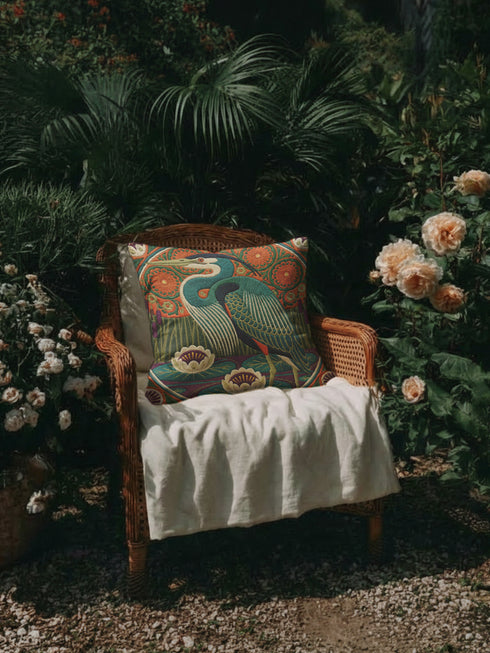 A square decorative pillowcase printed with a smooth, tapestry-like design. The central image is a large Great Blue Heron in profile, rendered in shades of teal, navy, and cream. The bird stands among stylized water lilies with cream petals and mustard centers, resting on deep plum and green waters. The background features tall green grass, purple cattails, and large, sun-like circular orange floral medallions. The detailed, embroidered-style pattern uses rich, earthy, maximalist colors.
