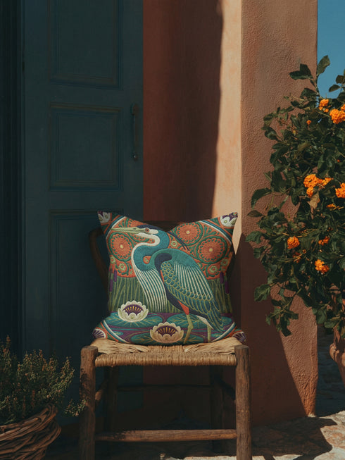 A square decorative pillowcase printed with a smooth, tapestry-like design. The central image is a large Great Blue Heron in profile, rendered in shades of teal, navy, and cream. The bird stands among stylized water lilies with cream petals and mustard centers, resting on deep plum and green waters. The background features tall green grass, purple cattails, and large, sun-like circular orange floral medallions. The detailed, embroidered-style pattern uses rich, earthy, maximalist colors.