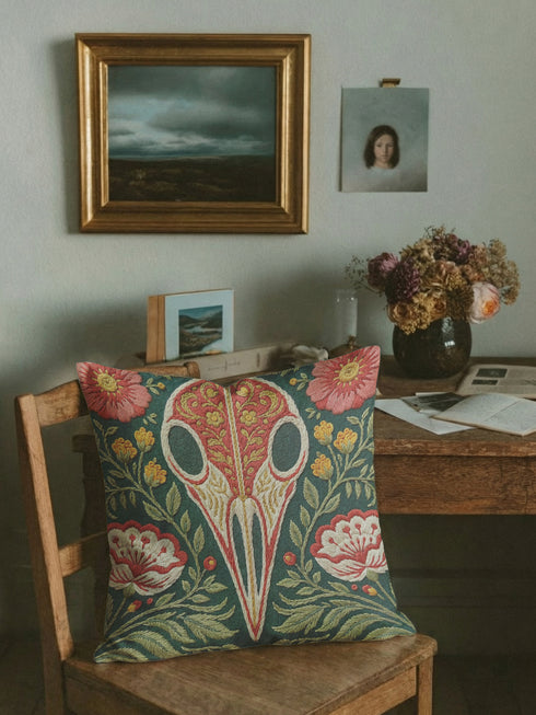 Square decorative pillow cover with a deep teal-green ground and a symmetrical folk-botanical design rendered in a faux embroidery style. At the center is a raven skull in cream and ivory, outlined in coral red, its cranium filled with golden scrolling filigree. Coral-pink roses sit in the upper corners, with red and white ruffled flowers, clustered yellow blooms, red berries, and green ferns framing the skull. The printed satin-stitch detailing mimics dense hand embroidery across the cover.
