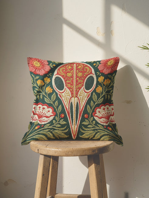 Square decorative pillow cover with a deep teal-green ground and a symmetrical folk-botanical design rendered in a faux embroidery style. At the center is a raven skull in cream and ivory, outlined in coral red, its cranium filled with golden scrolling filigree. Coral-pink roses sit in the upper corners, with red and white ruffled flowers, clustered yellow blooms, red berries, and green ferns framing the skull. The printed satin-stitch detailing mimics dense hand embroidery across the cover.