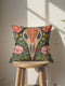 Square decorative pillow cover with a deep teal-green ground and a symmetrical folk-botanical design rendered in a faux embroidery style. At the center is a raven skull in cream and ivory, outlined in coral red, its cranium filled with golden scrolling filigree. Coral-pink roses sit in the upper corners, with red and white ruffled flowers, clustered yellow blooms, red berries, and green ferns framing the skull. The printed satin-stitch detailing mimics dense hand embroidery across the cover.