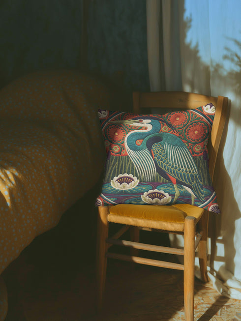 A square decorative pillowcase printed with a smooth, tapestry-like design. The central image is a large Great Blue Heron in profile, rendered in shades of teal, navy, and cream. The bird stands among stylized water lilies with cream petals and mustard centers, resting on deep plum and green waters. The background features tall green grass, purple cattails, and large, sun-like circular orange floral medallions. The detailed, embroidered-style pattern uses rich, earthy, maximalist colors.