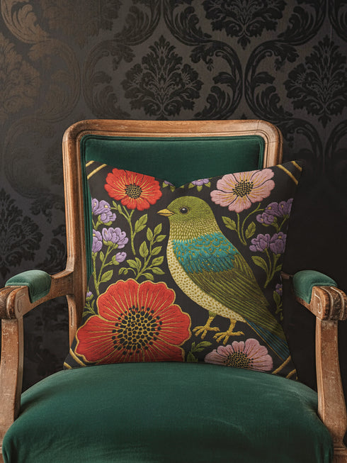 Square decorative pillow cover with a near-black ground and a richly colored Art Nouveau botanical design rendered in a faux embroidery style. At the center perches a songbird with an olive and chartreuse body, a teal wing patch, and golden-yellow beak and feet. Around it bloom large red-orange poppies with golden centers, clusters of lilac and lavender flowers, and pale pink daisy-like blossoms among green leaves. The printed satin-stitch detailing mimics dense hand embroidery across the cover.