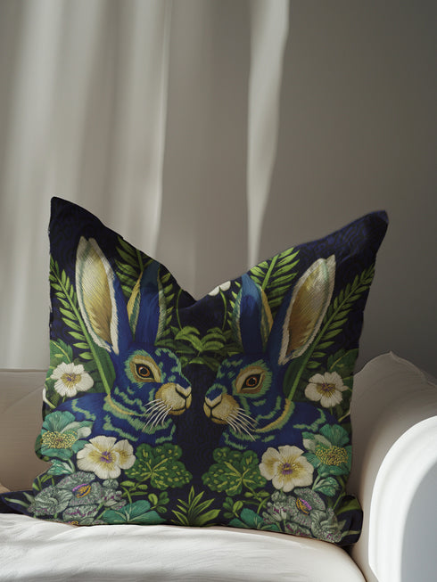 A Grimoire and Goblet decorative throw pillow cover featuring a high-fidelity print of two symmetrical hares facing each other. The design mimics the texture of raised embroidery with faux-stitches in antique gold, ochre, and royal blue threads against a deep midnight indigo background. The rabbits are surrounded by a lush wreath of green ferns and cream primrose flowers in a vintage medallion style. The fabric has a smooth, linen-like texture, visible as a soft matte finish