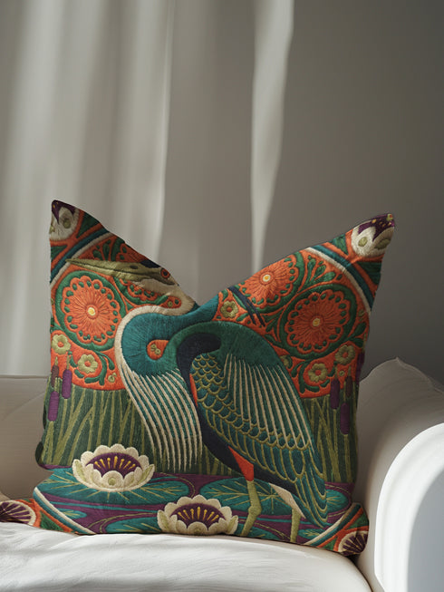 A square decorative pillowcase printed with a smooth, tapestry-like design. The central image is a large Great Blue Heron in profile, rendered in shades of teal, navy, and cream. The bird stands among stylized water lilies with cream petals and mustard centers, resting on deep plum and green waters. The background features tall green grass, purple cattails, and large, sun-like circular orange floral medallions. The detailed, embroidered-style pattern uses rich, earthy, maximalist colors.