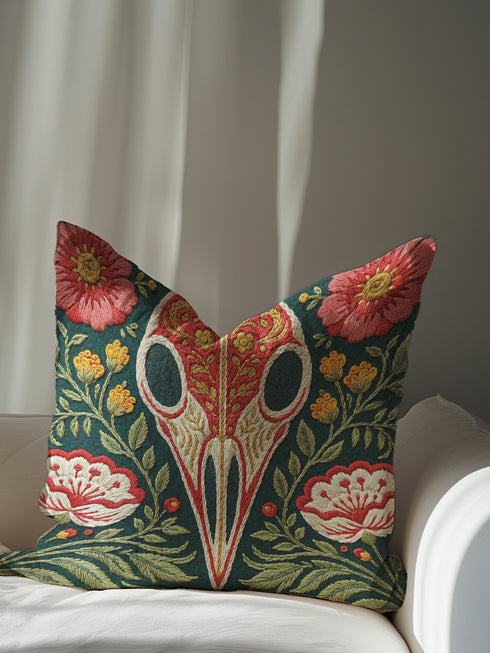 Square decorative pillow cover with a deep teal-green ground and a symmetrical folk-botanical design rendered in a faux embroidery style. At the center is a raven skull in cream and ivory, outlined in coral red, its cranium filled with golden scrolling filigree. Coral-pink roses sit in the upper corners, with red and white ruffled flowers, clustered yellow blooms, red berries, and green ferns framing the skull. The printed satin-stitch detailing mimics dense hand embroidery across the cover.