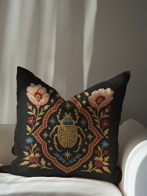 A square, dark charcoal pillow cover with a linen-feel texture, featuring an intricate, embroidered-style design. At the center, a large Egyptian scarab beetle in gold and turquoise is framed by a red and gold quatrefoil border. Symmetrical blush-pink poppies and smaller blue and red flowers with golden foliage flank the central motif, creating a lush botanical scene. Small celestial stars in gold and blue are scattered, adding a mystical, celestial element to this piece of dark academia decor.