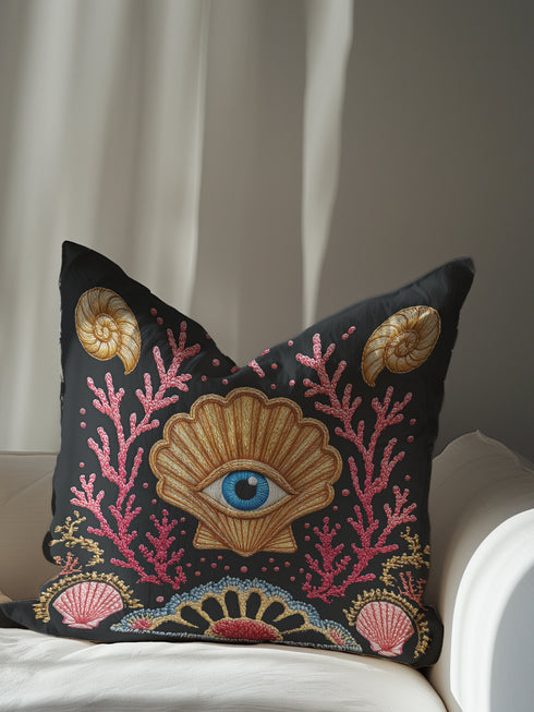 A square decorative pillow cover in a dark charcoal gray with a linen-feel texture. The design features a surrealist, mystical, and nautical goth motif. In the center is a large, golden scallop shell containing a realistic blue and white human eye, serving as an all-seeing eye or protection talisman. The shell is flanked by intricate, embroidered-style pink and red coral branches. The design is bordered by ammonite fossils and smaller seashells, creating dark academia and sea witch aesthetic.