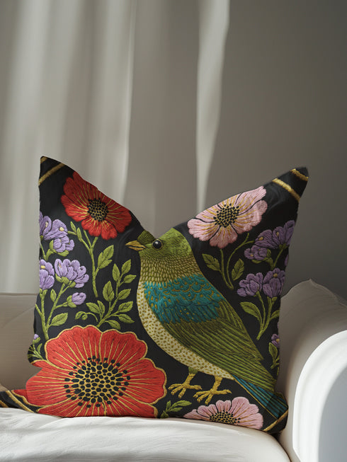 Square decorative pillow cover with a near-black ground and a richly colored Art Nouveau botanical design rendered in a faux embroidery style. At the center perches a songbird with an olive and chartreuse body, a teal wing patch, and golden-yellow beak and feet. Around it bloom large red-orange poppies with golden centers, clusters of lilac and lavender flowers, and pale pink daisy-like blossoms among green leaves. The printed satin-stitch detailing mimics dense hand embroidery across the cover.