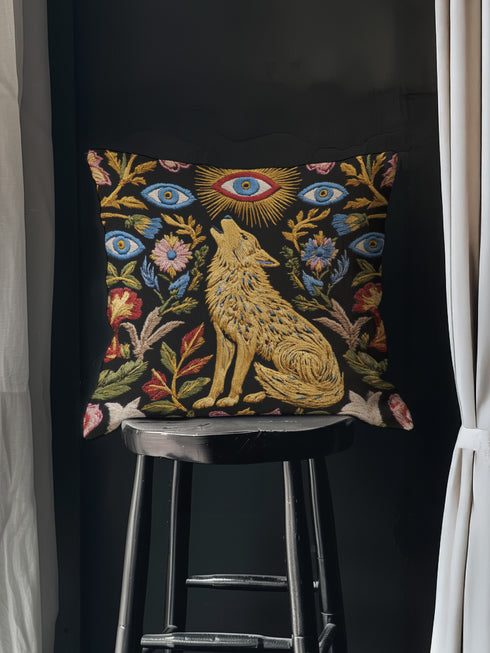 A square decorative throw pillow case with a dark charcoal background and a detailed, faux embroidery design. In the center, a golden-yellow howling wolf sits among colorful botanical flowers and leaves. Above the wolf is a celestial sunburst with a single protective evil eye. The central design is framed by more floral elements and six additional blue evil eye motifs, creating a symmetrical, mystical pattern. This gothic, witchy cushion cover has a distinct dark academia and bohemian aesthetic.