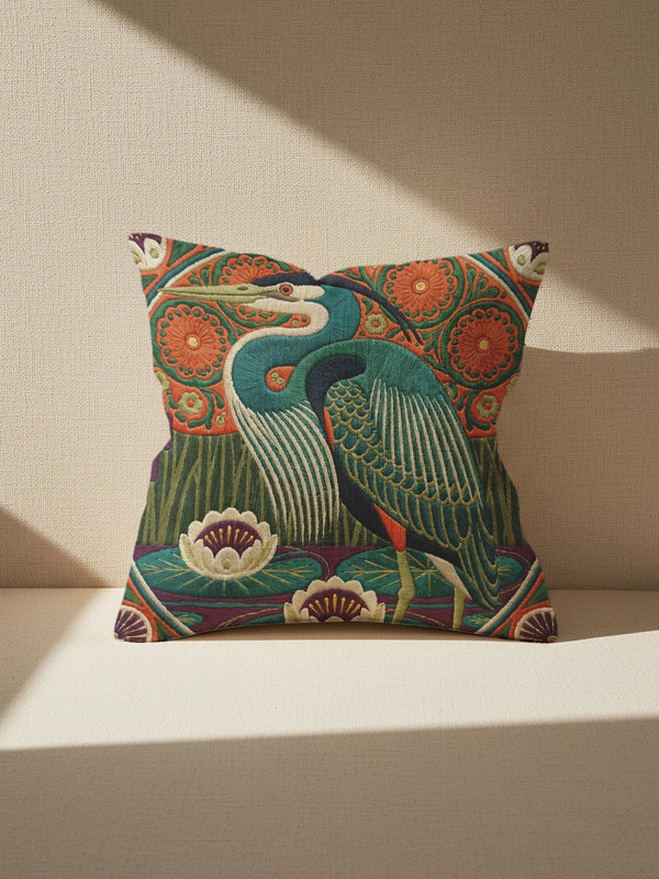 A square decorative pillowcase printed with a smooth, tapestry-like design. The central image is a large Great Blue Heron in profile, rendered in shades of teal, navy, and cream. The bird stands among stylized water lilies with cream petals and mustard centers, resting on deep plum and green waters. The background features tall green grass, purple cattails, and large, sun-like circular orange floral medallions. The detailed, embroidered-style pattern uses rich, earthy, maximalist colors.
