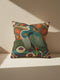 A square decorative pillowcase printed with a smooth, tapestry-like design. The central image is a large Great Blue Heron in profile, rendered in shades of teal, navy, and cream. The bird stands among stylized water lilies with cream petals and mustard centers, resting on deep plum and green waters. The background features tall green grass, purple cattails, and large, sun-like circular orange floral medallions. The detailed, embroidered-style pattern uses rich, earthy, maximalist colors.