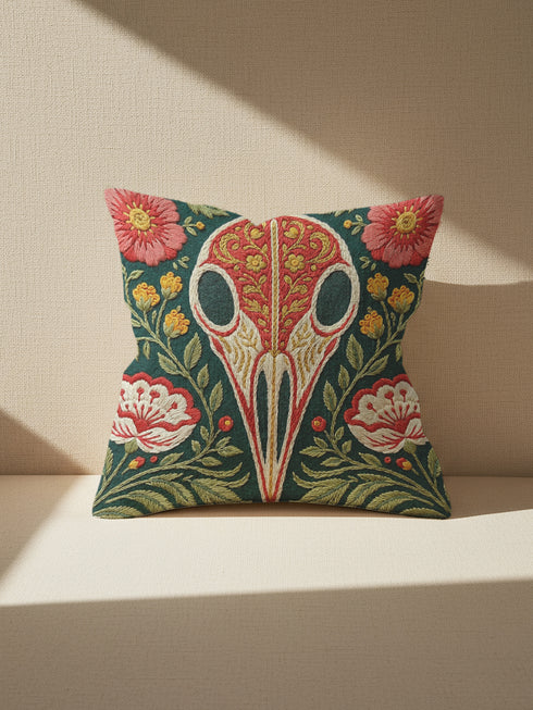 Square decorative pillow cover with a deep teal-green ground and a symmetrical folk-botanical design rendered in a faux embroidery style. At the center is a raven skull in cream and ivory, outlined in coral red, its cranium filled with golden scrolling filigree. Coral-pink roses sit in the upper corners, with red and white ruffled flowers, clustered yellow blooms, red berries, and green ferns framing the skull. The printed satin-stitch detailing mimics dense hand embroidery across the cover.