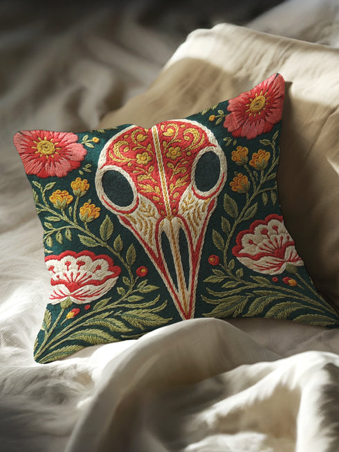 Square decorative pillow cover with a deep teal-green ground and a symmetrical folk-botanical design rendered in a faux embroidery style. At the center is a raven skull in cream and ivory, outlined in coral red, its cranium filled with golden scrolling filigree. Coral-pink roses sit in the upper corners, with red and white ruffled flowers, clustered yellow blooms, red berries, and green ferns framing the skull. The printed satin-stitch detailing mimics dense hand embroidery across the cover.