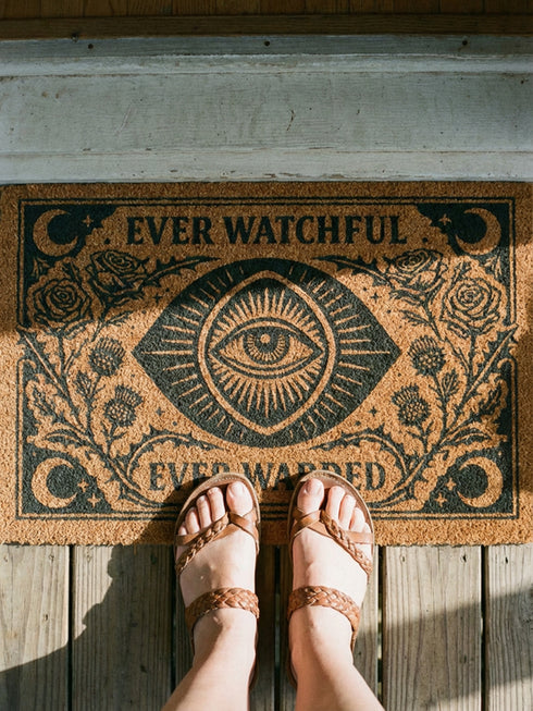 A premium 24x16 inch Grimoire and Goblet Warding Entry Mat featuring a centralized All-Seeing Eye medallion in high-contrast black vinyl on natural tan coconut coir. The design includes radial sunbursts, crescent moons, stars, and botanical roses and thistles. Serif text reads EVER WATCHFUL and EVER WARDED. The rug features a heavy-duty black vinyl backing for weather-resistant stability. Perfect for a luxury Victorian Gothic or Dark Academia entryway sanctuary.