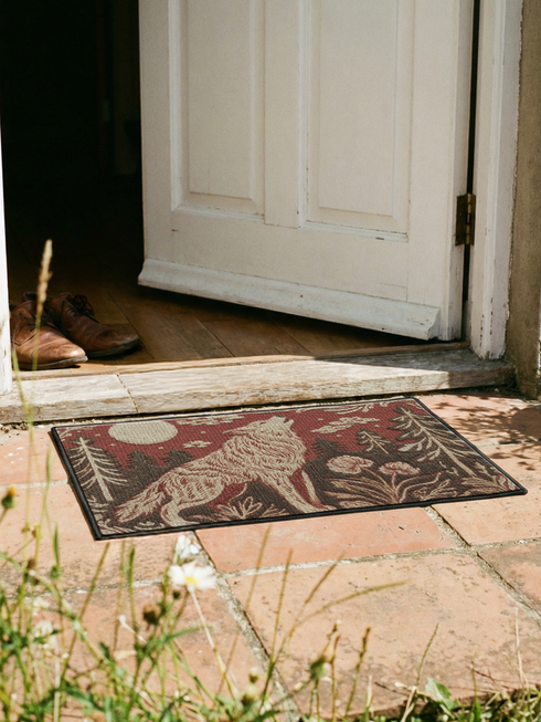 A rectangular Grimoire and Goblet doormat featuring a howling wolf standing in a dark forest under a full cream moon. The design utilizes a rust red and deep brown color palette with a high-fidelity 2D print technique that mimics the texture of vintage tapestry needlepoint. Stylized pine trees and forest flora surround the wolf. The mat has black trim and is made of smooth polyester, creating a rustic cottagecore aesthetic without actual raised threads or embroidery.