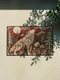 A rectangular Grimoire and Goblet doormat featuring a howling wolf standing in a dark forest under a full cream moon. The design utilizes a rust red and deep brown color palette with a high-fidelity 2D print technique that mimics the texture of vintage tapestry needlepoint. Stylized pine trees and forest flora surround the wolf. The mat has black trim and is made of smooth polyester, creating a rustic cottagecore aesthetic without actual raised threads or embroidery.