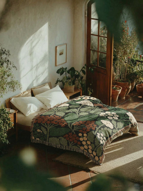 A Grimoire and Goblet luxury woven throw blanket featuring a dark botanical design. The Jacquard cotton tapestry showcases cream elderflower clusters and rust-colored berries against a deep black background. Sage green leaves interweave throughout the floral pattern. The blanket is finished with multicolored fringe edges. A moody cottagecore aesthetic piece ideal for dark academia decor.