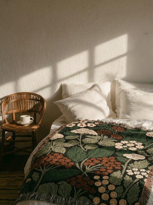 A Grimoire and Goblet luxury woven throw blanket featuring a dark botanical design. The Jacquard cotton tapestry showcases cream elderflower clusters and rust-colored berries against a deep black background. Sage green leaves interweave throughout the floral pattern. The blanket is finished with multicolored fringe edges. A moody cottagecore aesthetic piece ideal for dark academia decor.