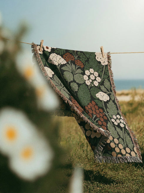 A Grimoire and Goblet luxury woven throw blanket featuring a dark botanical design. The Jacquard cotton tapestry showcases cream elderflower clusters and rust-colored berries against a deep black background. Sage green leaves interweave throughout the floral pattern. The blanket is finished with multicolored fringe edges. A moody cottagecore aesthetic piece ideal for dark academia decor.