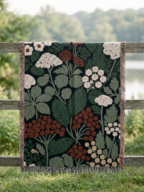 A Grimoire and Goblet luxury woven throw blanket featuring a dark botanical design. The Jacquard cotton tapestry showcases cream elderflower clusters and rust-colored berries against a deep black background. Sage green leaves interweave throughout the floral pattern. The blanket is finished with multicolored fringe edges. A moody cottagecore aesthetic piece ideal for dark academia decor.