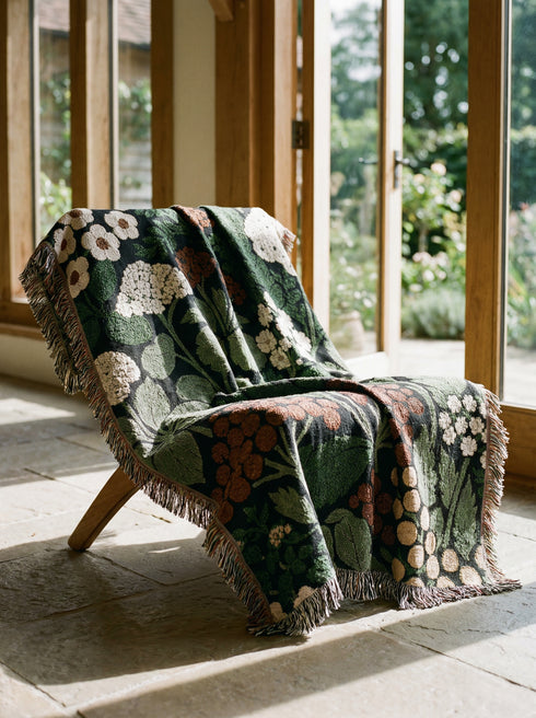 A Grimoire and Goblet luxury woven throw blanket featuring a dark botanical design. The Jacquard cotton tapestry showcases cream elderflower clusters and rust-colored berries against a deep black background. Sage green leaves interweave throughout the floral pattern. The blanket is finished with multicolored fringe edges. A moody cottagecore aesthetic piece ideal for dark academia decor.