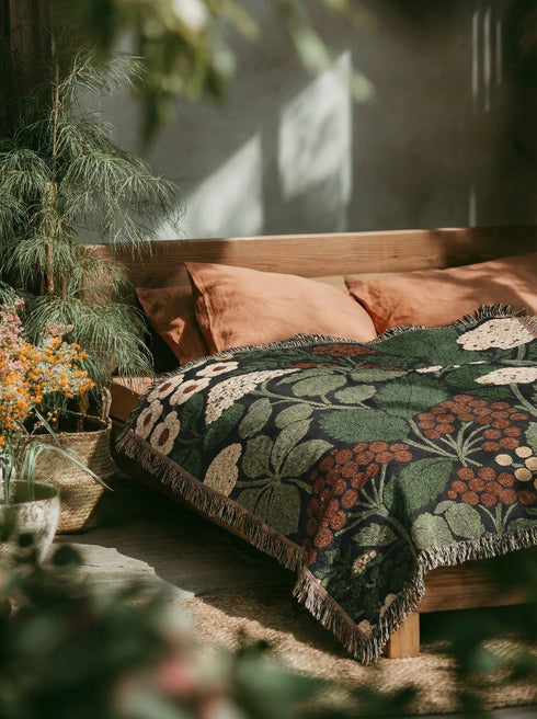 A Grimoire and Goblet luxury woven throw blanket featuring a dark botanical design. The Jacquard cotton tapestry showcases cream elderflower clusters and rust-colored berries against a deep black background. Sage green leaves interweave throughout the floral pattern. The blanket is finished with multicolored fringe edges. A moody cottagecore aesthetic piece ideal for dark academia decor.