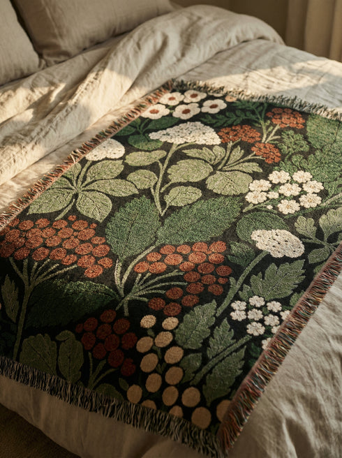 A Grimoire and Goblet luxury woven throw blanket featuring a dark botanical design. The Jacquard cotton tapestry showcases cream elderflower clusters and rust-colored berries against a deep black background. Sage green leaves interweave throughout the floral pattern. The blanket is finished with multicolored fringe edges. A moody cottagecore aesthetic piece ideal for dark academia decor.