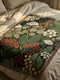 A Grimoire and Goblet luxury woven throw blanket featuring a dark botanical design. The Jacquard cotton tapestry showcases cream elderflower clusters and rust-colored berries against a deep black background. Sage green leaves interweave throughout the floral pattern. The blanket is finished with multicolored fringe edges. A moody cottagecore aesthetic piece ideal for dark academia decor.