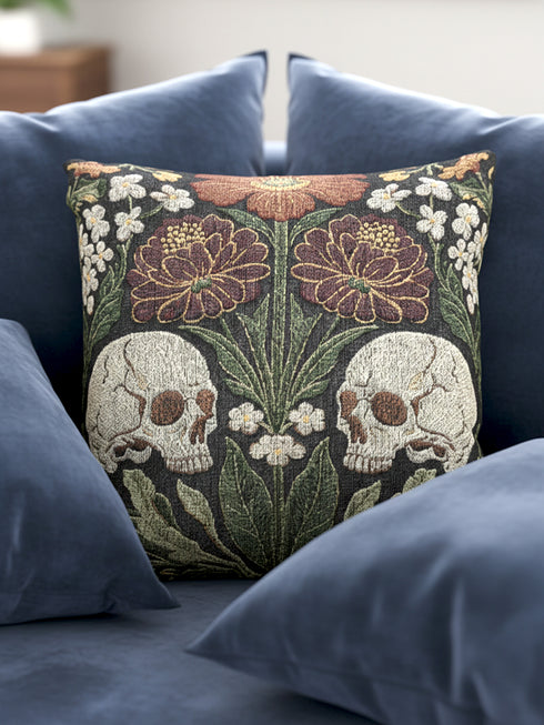 A Grimoire and Goblet woven tapestry pillow featuring a symmetrical dark botanical design. Two beige human skulls face each other in profile against a dark charcoal background. They are surrounded by deep burgundy dahlia flowers, sage green leaves, and small white floral clusters in an Arts and Crafts style. The texture is a visible fabric weave. The design is double-sided, square, and filled.