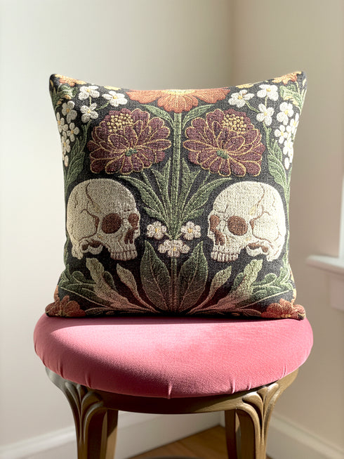 A Grimoire and Goblet woven tapestry pillow featuring a symmetrical dark botanical design. Two beige human skulls face each other in profile against a dark charcoal background. They are surrounded by deep burgundy dahlia flowers, sage green leaves, and small white floral clusters in an Arts and Crafts style. The texture is a visible fabric weave. The design is double-sided, square, and filled.