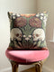 A Grimoire and Goblet woven tapestry pillow featuring a symmetrical dark botanical design. Two beige human skulls face each other in profile against a dark charcoal background. They are surrounded by deep burgundy dahlia flowers, sage green leaves, and small white floral clusters in an Arts and Crafts style. The texture is a visible fabric weave. The design is double-sided, square, and filled.