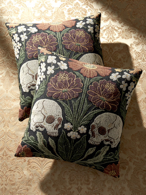 A Grimoire and Goblet woven tapestry pillow featuring a symmetrical dark botanical design. Two beige human skulls face each other in profile against a dark charcoal background. They are surrounded by deep burgundy dahlia flowers, sage green leaves, and small white floral clusters in an Arts and Crafts style. The texture is a visible fabric weave. The design is double-sided, square, and filled.