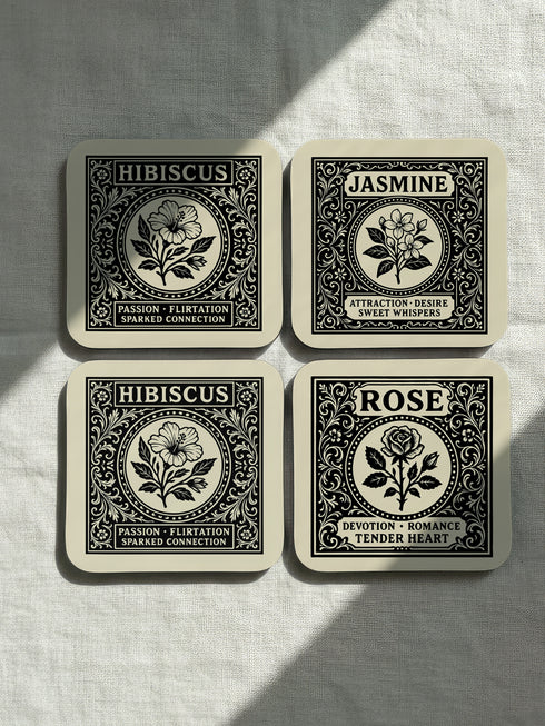 A set of four glossy square coasters by Grimoire and Goblet, arranged on a surface. Each coaster features a white Art Nouveau botanical illustration on a black background with text. The designs are: Rose "Devotion Romance," Hawthorn "Heart Guardian," Hibiscus "Passion Flirtation," and Jasmine "Attraction Desire." The style is Dark Academia and Victorian Gothic. The high-gloss finish reflects light, highlighting the intricate floral details and cork backing.