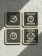 A set of four glossy square coasters by Grimoire and Goblet, arranged on a surface. Each coaster features a white Art Nouveau botanical illustration on a black background with text. The designs are: Rose "Devotion Romance," Hawthorn "Heart Guardian," Hibiscus "Passion Flirtation," and Jasmine "Attraction Desire." The style is Dark Academia and Victorian Gothic. The high-gloss finish reflects light, highlighting the intricate floral details and cork backing.