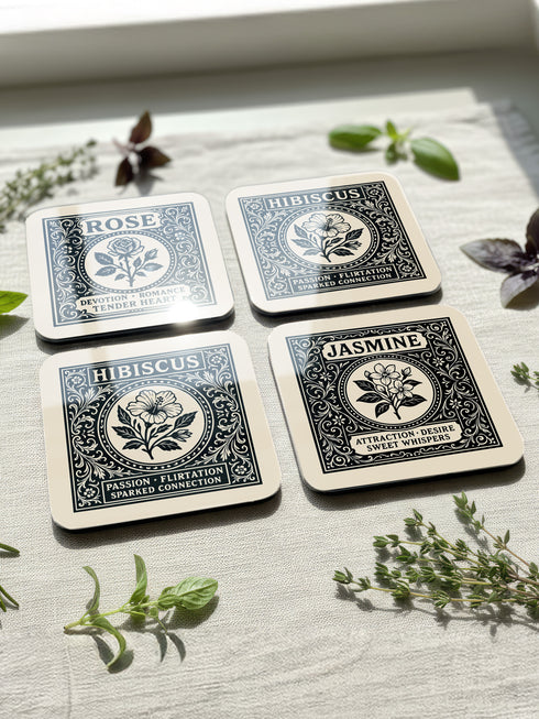 A set of four glossy square coasters by Grimoire and Goblet, arranged on a surface. Each coaster features a white Art Nouveau botanical illustration on a black background with text. The designs are: Rose "Devotion Romance," Hawthorn "Heart Guardian," Hibiscus "Passion Flirtation," and Jasmine "Attraction Desire." The style is Dark Academia and Victorian Gothic. The high-gloss finish reflects light, highlighting the intricate floral details and cork backing.