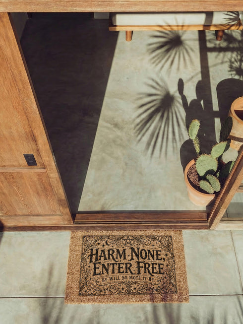 A rectangular natural coir doormat featuring the Wiccan Rede "An' It Harm None, Do What Ye Will" in a gothic dark academia style. The central typography is surrounded by symmetrical botanical flourishes and mystical symbols printed in high-contrast black ink. The 2D flat print mimics the look of a vintage grimoire page on the coarse, tan coconut fiber surface. The mat has a durable black vinyl backing and is pictured at a front door as a protective threshold guardian.