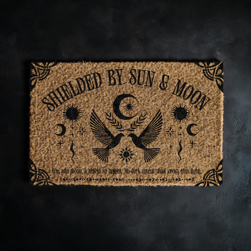 Natural coir doormat featuring two symmetrical doves beneath a crescent moon, surrounded by celestial and botanical accents. The phrase Shielded by Sun & Moon is stenciled above, with a protective incantation at the base. Non-slip vinyl backing for outdoor use.