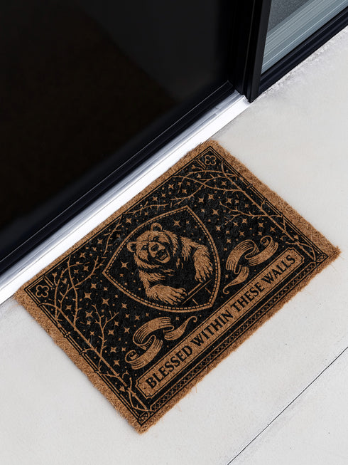 A rectangular eco-friendly coir doormat measuring 24 by 16 inches displayed on a stone porch. The natural golden-brown coconut fiber mat features a bold black graphic design of a heraldic bear head centered on a medieval knight's shield. Above and below the shield, Gothic-style typography reads BLESSED WITHIN THESE WALLS. The mat has a durable black vinyl non-slip backing. Ideal for Castlecore, Dark Academia, and Grimoire and Goblet style gothic home decor themes.