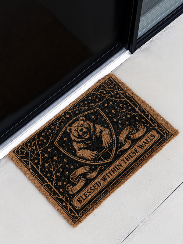A rectangular eco-friendly coir doormat measuring 24 by 16 inches displayed on a stone porch. The natural golden-brown coconut fiber mat features a bold black graphic design of a heraldic bear head centered on a medieval knight's shield. Above and below the shield, Gothic-style typography reads BLESSED WITHIN THESE WALLS. The mat has a durable black vinyl non-slip backing. Ideal for Castlecore, Dark Academia, and Grimoire and Goblet style gothic home decor themes.