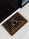 A rectangular eco-friendly coir doormat measuring 24 by 16 inches displayed on a stone porch. The natural golden-brown coconut fiber mat features a bold black graphic design of a heraldic bear head centered on a medieval knight's shield. Above and below the shield, Gothic-style typography reads BLESSED WITHIN THESE WALLS. The mat has a durable black vinyl non-slip backing. Ideal for Castlecore, Dark Academia, and Grimoire and Goblet style gothic home decor themes.