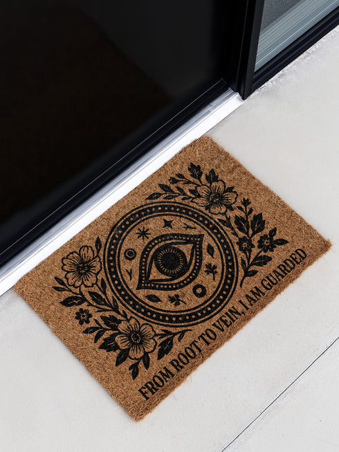 Rectangular 24x16 inch coconut coir doormat with a black vinyl backing. The mat features a central evil eye surrounded by botanical flowers, stars, and dots, with the phrase “From Root to Vein, I Am Guarded” printed at the bottom. Designed for outdoor use, with durable bristle texture for entryways or garden paths.